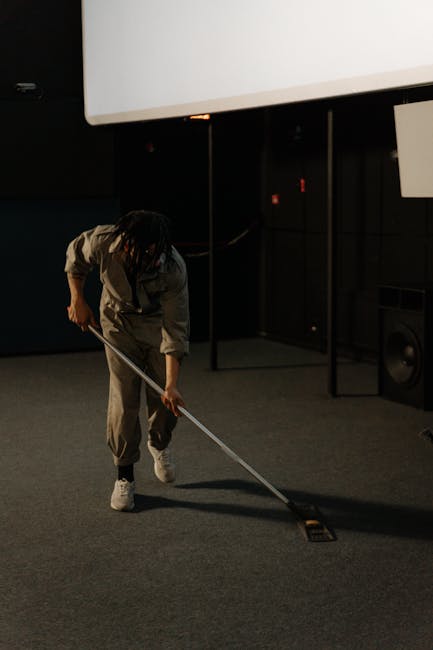 Close-up image of a person vacuuming a traditional patterned area rug with a modern handheld vacuum cleaner, revealing the detailed floral design in shades of beige, green, and orange. The scene is il